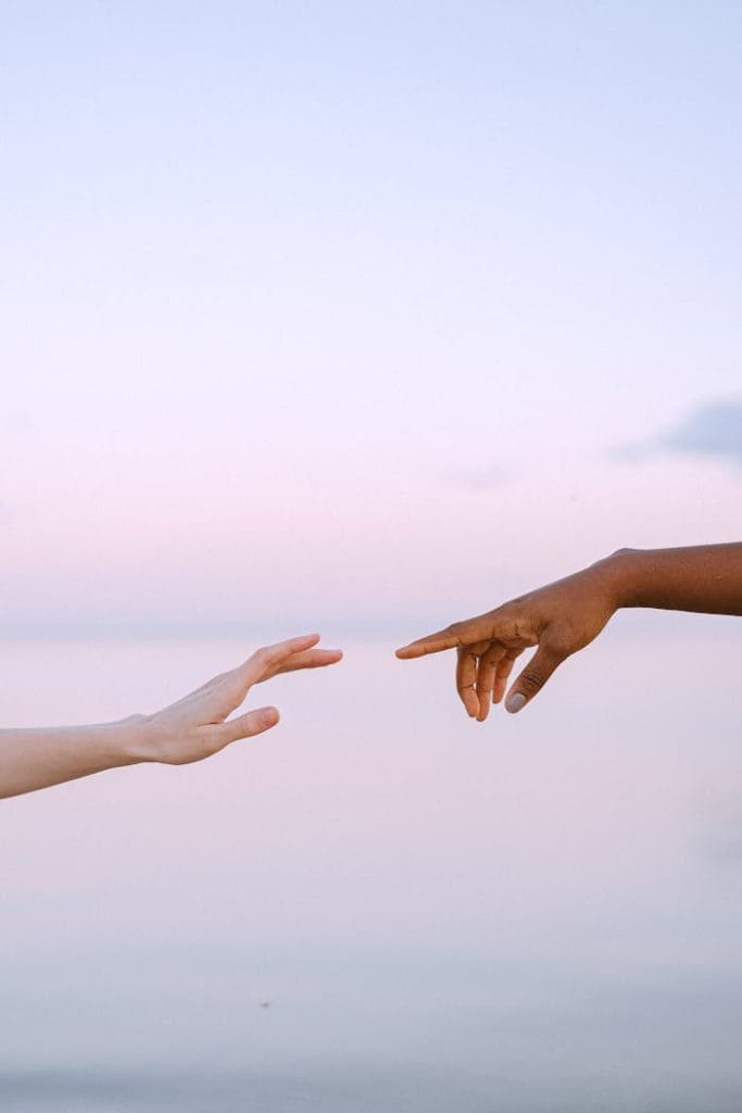 photo-of-people-reaching-each-other-s-hands-4672714 Two diverse hands reaching towards each other over a serene pastel sky and water background.