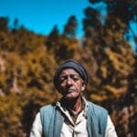 A senior man wearing a beanie stands outdoors against a blurred forest background.